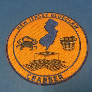 May include: Orange and blue embroidered patch with the text "New Jersey Blueclaw Crabber". The patch features a crab, a bucket, a crab trap, and an outline of the state of New Jersey.