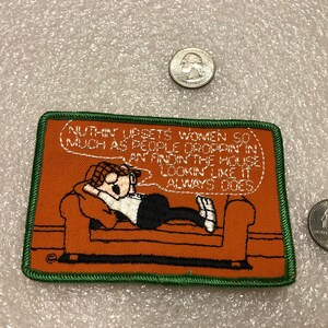May include: A green and orange embroidered patch with a cartoon man lying on a couch. The text on the patch reads: "NUTHIN' UPSETS WOMEN SO MUCH AS PEOPLE DROPPIN IN AN FINDIN THE HOUSE LOOKIN LIKE IT ALWAYS DOES."