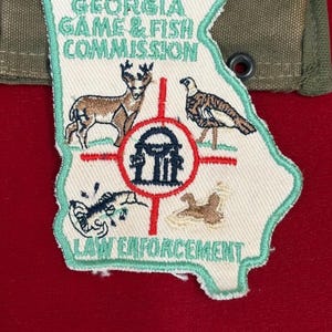 Puede incluir: Parche bordado con la forma del estado de Georgia, con ilustraciones de un ciervo, un pavo y otros animales salvajes. El parche tiene el texto "Georgia Game & Fish Commission Law Enforcement" en verde y blanco.