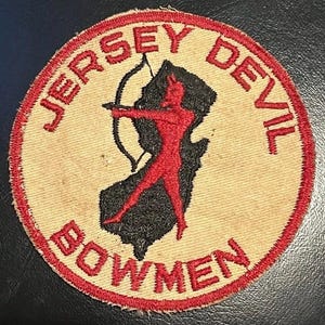 Könnte beinhalten: Runder Aufnäher mit rotem gesticktem Text und Rand. Der Text lautet "JERSEY DEVIL BOWMEN". Eine rote Teufelsfigur mit Pfeil und Bogen ist auf einer schwarzen Umrisslinie des Bundesstaates New Jersey zentriert.