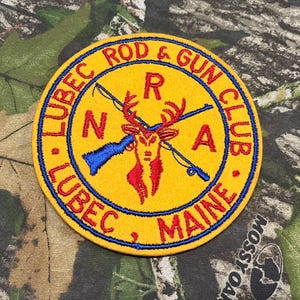 Può includere: Toppa circolare gialla con bordi blu e rossi. Presenta la scritta "LUBEC ROD & GUN CLUB, MAINE" in un motivo circolare. Un cervo con fucili e canne da pesca incrociate è al centro.
