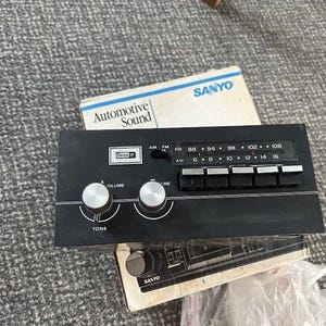 Peut inclure: Une radio de voiture Sanyo noire avec des boutons argentés pour le volume et la tonalité, ainsi qu'une série de boutons et d'indicateurs de fréquence. La radio est accompagnée d'une boîte intitulée "Automotive Sound".