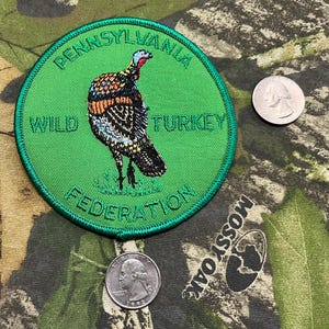 May include: A circular green patch with the text "PENNSYLVANIA WILD TURKEY FEDERATION" surrounds a detailed illustration of a wild turkey. Two quarters are next to the patch on a camouflage background.