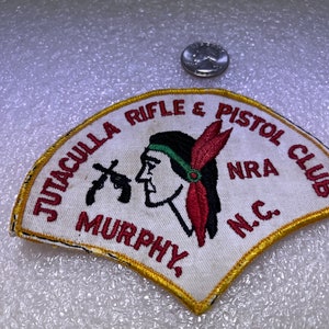 Op de afbeelding: Witte geborduurde patch met een gele rand. De patch toont een profiel van een inheemse Amerikaan met een rode verenhoed, twee gekruiste geweren en de tekst "Jutaculla Rifle & Pistol Club NRA N.C. Murphy."