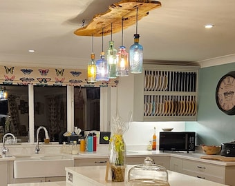 May include: A rustic wooden light fixture with five glass bottles hanging from it. The bottles are different colors and have a vintage look. The fixture is hanging over a kitchen counter with white cabinets.
