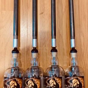 Puede incluir: Cuatro botellas de vidrio transparente con etiquetas negras y una ilustración de dibujos animados de un hombre barbudo. Las botellas están etiquetadas como "Big Peat" y tienen un contenido de alcohol del 46%. Las botellas están unidas a tuberías metálicas con tapas plateadas.