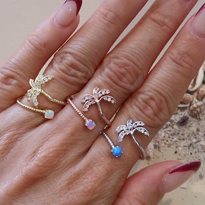 May include: A hand wearing four palm tree rings with different colored gemstones. The rings are on the index, middle, and ring fingers. The nails are painted with a burgundy french manicure.