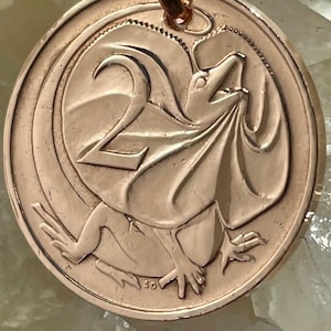 May include: A circular copper-toned pendant featuring a raised relief design of a frilled-neck lizard and the number 2. The pendant is attached to a small metal jump ring at the top, showcasing a metallic finish with detailed embossed textures.