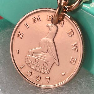 Puede incluir: Un llavero dorado con una moneda de Zimbabue de 1997. La moneda muestra un emblema de águila y la palabra "ZIMBABWE" en el borde. El llavero tiene un diseño de eslabones de cadena y un anillo dorado.