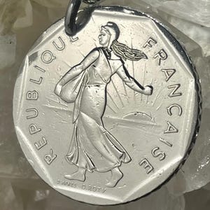 Könnte beinhalten: Ein silberfarbener, runder Anhänger mit dem Semeuse-Motiv, das eine schreitende Figur beim Säen vor einem aufgehenden Sonnenhintergrund zeigt. Der Text REPUBLIQUE FRANCAISE ist am Rand eingeprägt, mit O. ROTY am unteren Rand.
