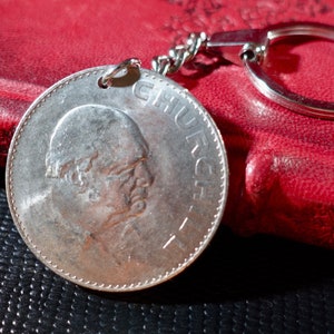 Peut inclure: Un porte-clés argenté avec un charme rond en forme de pièce de monnaie représentant le profil d'un homme et le mot "CHURCH" inscrit sur le côté. Le porte-clés est attaché à un carnet en cuir rouge.