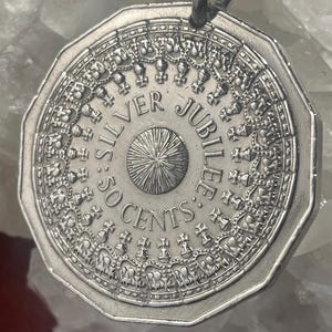 May include: A silver-coloured, dodecagonal coin pendant featuring a central sunburst design surrounded by the text SILVER JUBILEE 50 CENTS. The border is decorated with a repeating pattern of small crowns and crosses, with a metal loop at the top.