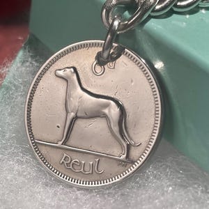 Puede incluir: Colgante de chapa para perro plateado con una silueta de galgo. El colgante est&aacute; sujeto a una cadena plateada. La palabra "Reul" est&aacute; inscrita en la parte inferior del colgante.