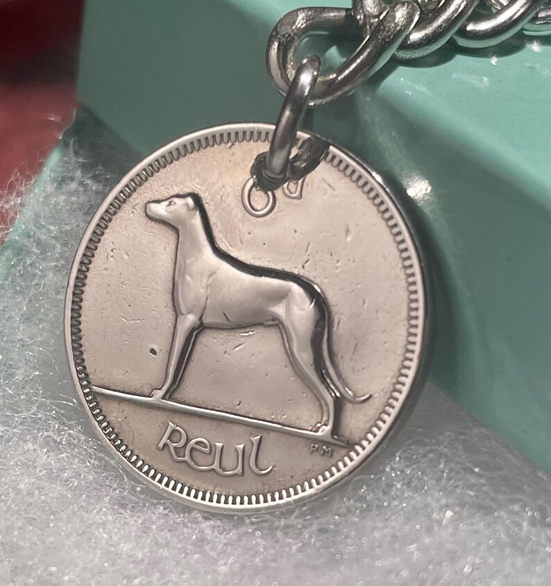 Puede incluir: Colgante de chapa para perro plateado con una silueta de galgo. El colgante est&aacute; sujeto a una cadena plateada. La palabra "Reul" est&aacute; inscrita en la parte inferior del colgante.