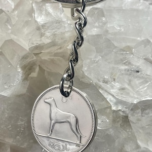Puede incluir: Llavero plateado con un colgante redondo con la silueta de un perro. El colgante est&aacute; unido a una cadena y un llavero plateados. La palabra "Reul" est&aacute; inscrita en el colgante. El fondo es una superficie clara y texturizada.