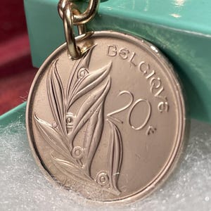May include: A silver-coloured metal keyring featuring a circular coin pendant. The coin displays a relief of lily of the valley flowers, the number 20, the letter F, and the text BELGIQUE. The pendant is attached to a metallic chain link.