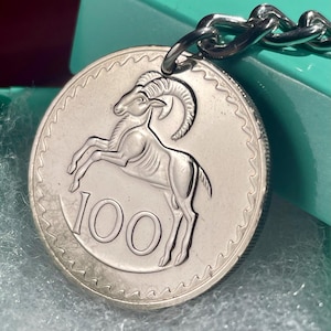 Pode incluir: Porta-chaves de moeda prateada com um relevo detalhado de um carneiro com grandes chifres curvos. A moeda exibe o número "100" e está presa a uma corrente prateada. O fundo é uma superfície de cor verde-azulado.