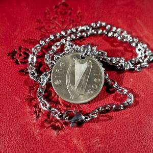 Puede incluir: Un collar plateado con un colgante de moneda que muestra un arpa y el a&ntilde;o 1969. La cadena es de eslabones con un cierre de mosquet&oacute;n. Descansa sobre una superficie roja texturizada.