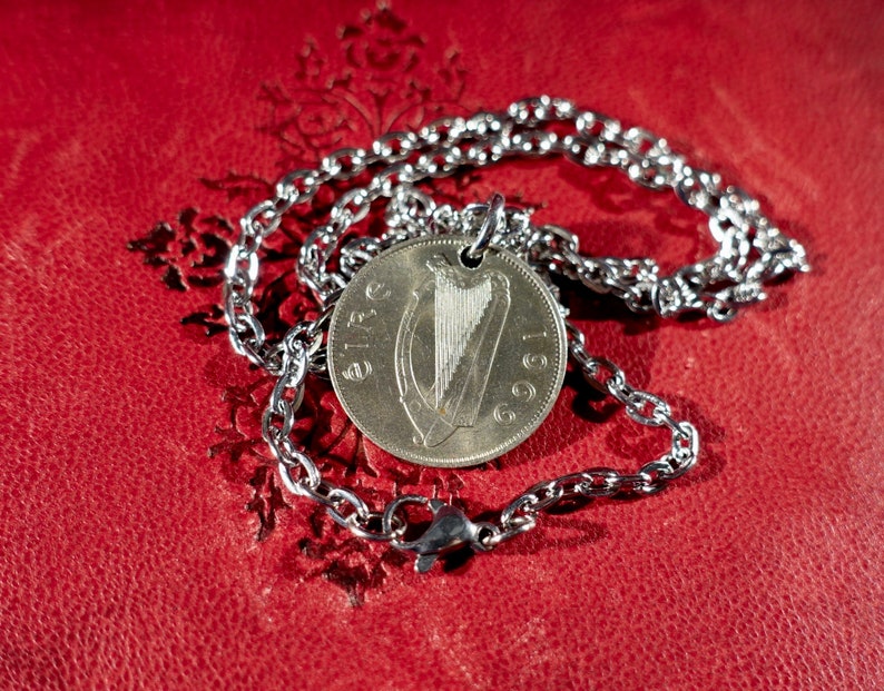 Puede incluir: Un collar plateado con un colgante de moneda que muestra un arpa y el a&ntilde;o 1969. La cadena es de eslabones con un cierre de mosquet&oacute;n. Descansa sobre una superficie roja texturizada.