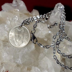 Puede incluir: Un collar plateado con un colgante de moneda. La moneda muestra un arpa y el a&ntilde;o 1969. La cadena est&aacute; hecha de peque&ntilde;as piezas de metal unidas. El fondo es una superficie clara y texturizada.