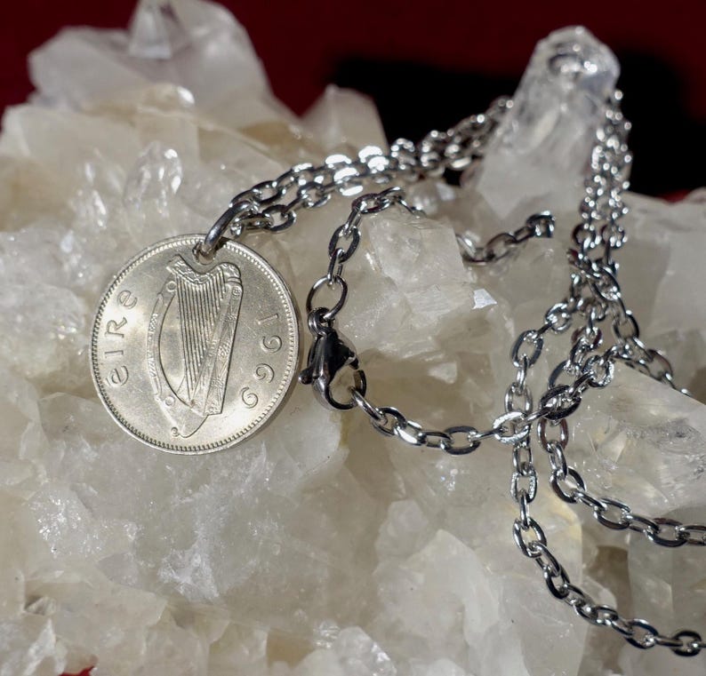 Puede incluir: Un collar plateado con un colgante de moneda. La moneda muestra un arpa y el a&ntilde;o 1969. La cadena est&aacute; hecha de peque&ntilde;as piezas de metal unidas. El fondo es una superficie clara y texturizada.