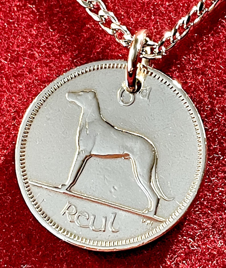 Puede incluir: Collar con colgante plateado. El colgante redondo presenta la silueta de un perro y la palabra "Reul". El colgante cuelga de una fina cadena plateada. El fondo es rojo oscuro.