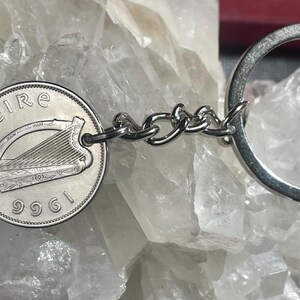 Puede incluir: Un llavero plateado con una moneda irlandesa de 1961. La moneda muestra un arpa y la palabra "eire". El llavero est&aacute; conectado por una cadena de peque&ntilde;os eslabones plateados y un anillo plateado.