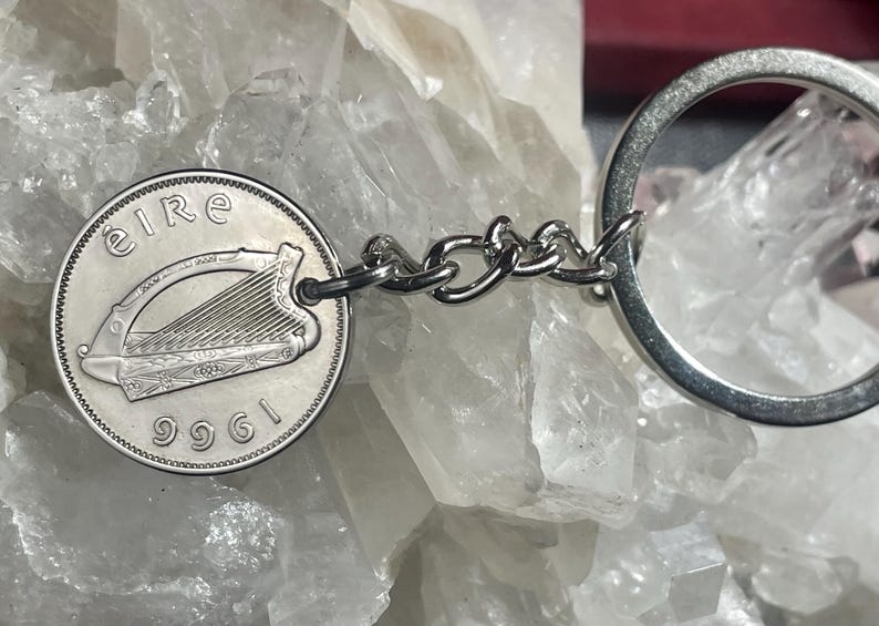 Puede incluir: Un llavero plateado con una moneda irlandesa de 1961. La moneda muestra un arpa y la palabra "eire". El llavero est&aacute; conectado por una cadena de peque&ntilde;os eslabones plateados y un anillo plateado.