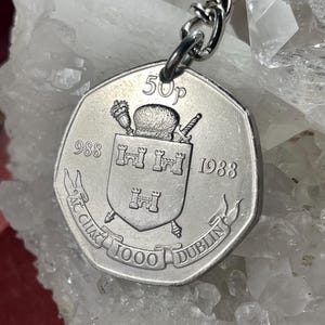 Puede incluir: Un llavero plateado con una moneda de 50p de 1988. La moneda muestra un escudo de armas con tres castillos y el texto "AC CHIACH 1000 DUBLIN". El llavero está sujeto a una cadena plateada.