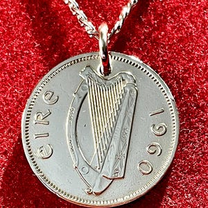 Puede incluir: Colgante de moneda de plata en una cadena de plata. La moneda presenta un dise&ntilde;o de arpa y el a&ntilde;o 1960. La palabra "Eire" est&aacute; inscrita en la moneda. El colgante cuelga sobre un fondo rojo.