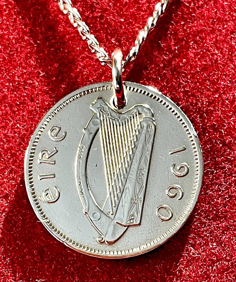 Puede incluir: Colgante de moneda de plata en una cadena de plata. La moneda presenta un dise&ntilde;o de arpa y el a&ntilde;o 1960. La palabra "Eire" est&aacute; inscrita en la moneda. El colgante cuelga sobre un fondo rojo.