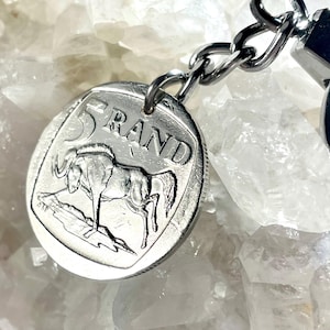 May include: A silver keychain featuring a South African 5 Rand coin with a depiction of a springbok antelope. The coin is attached to a silver chain with a silver ring.