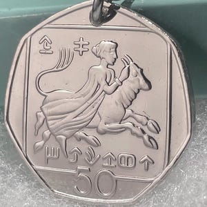 Könnte beinhalten: Ein silberfarbener, siebeneckiger Münzanhänger mit einem geprägten Motiv einer Figur, die auf einer Ziege reitet. Die Münze zeigt unten die Zahl 50 sowie verschiedene stilisierte Symbole und Zeichen, die über und unter der zentralen Figur angeordnet sind.
