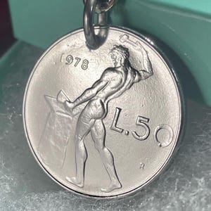 May include: Silver-colored coin pendant with a detailed relief of a muscular figure hammering metal, dated 1978. The coin reads "L.50" and is attached to a silver-tone chain, suitable for a necklace or keychain.