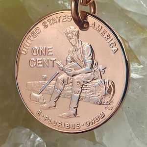 May include: A copper-colored pendant featuring a United States one-cent coin. The coin depicts a seated figure reading a book, with the words "UNITED STATES OF AMERICA" and "ONE CENT" visible. The pendant is attached to a rose gold chain.