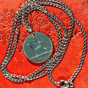 Puede incluir: Collar plateado con un colgante circular que representa un galgo. El colgante cuelga de una fina cadena con un cierre de mosquet&oacute;n. El collar se muestra sobre un fondo rojo.