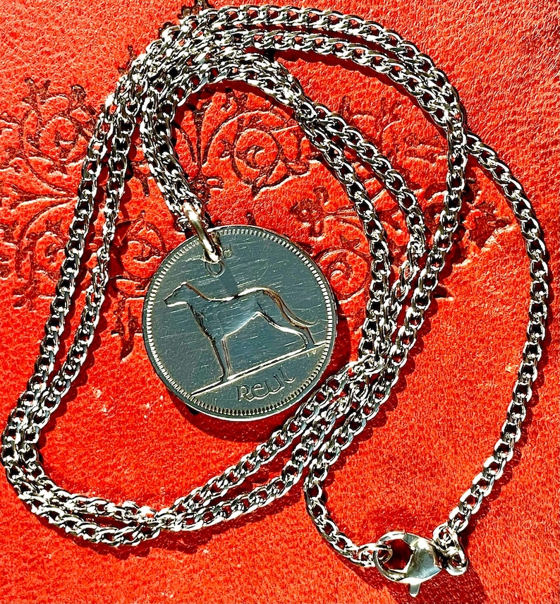 Puede incluir: Collar plateado con un colgante circular que representa un galgo. El colgante cuelga de una fina cadena con un cierre de mosquet&oacute;n. El collar se muestra sobre un fondo rojo.