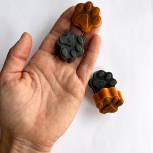 Könnte beinhalten: Vier pfotenförmige Wachsschmelzen in Grau- und Orangetönen. Die Pfotenabdrücke sind detailliert, und die Schmelzen sind auf einem weißen Hintergrund angeordnet. Die Schmelzen sind klein, und das Bild ist gut beleuchtet.