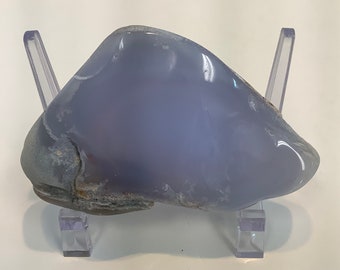 Può includere: Un grande ciottolo di agata levigato, traslucido e blu chiaro, su un supporto in plastica trasparente. Il ciottolo ha una superficie liscia e arrotondata e una base ruvida e testurizzata.