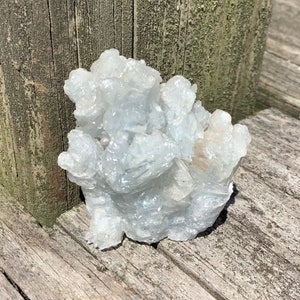 Pu&ograve; includere: Un gruppo di cristalli di celestite bianchi con un bagliore iridescente e scintillante. I cristalli sono disposti in una forma naturale e organica, che ricorda una piccola montagna.