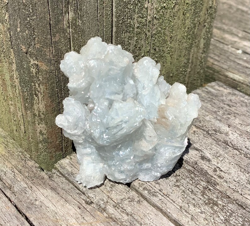 Pu&ograve; includere: Un gruppo di cristalli di celestite bianchi con un bagliore iridescente e scintillante. I cristalli sono disposti in una forma naturale e organica, che ricorda una piccola montagna.