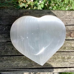 May include: A white selenite heart shaped crystal on a wooden surface. The crystal has a smooth, polished finish and is translucent.