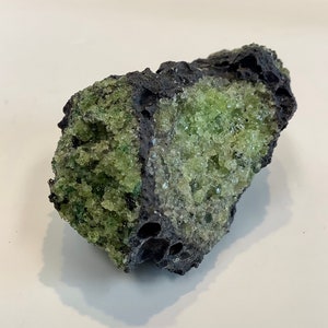 May include: A rough, dark gray rock with a large section of bright green crystals embedded in it. The crystals are clustered together and have a sparkly, almost glassy appearance.