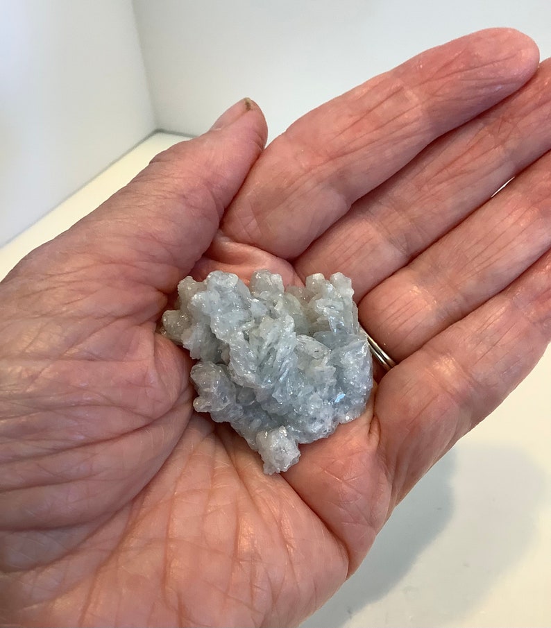 Pu&ograve; includere: Un ammasso di piccoli cristalli bianchi e blu chiaro in una mano. I cristalli sono raggruppati in una forma grezza e organica.