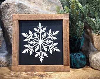 Peut inclure: Panneau carré en bois avec un flocon de neige blanc sur fond noir. Le panneau est encadré de bois brun foncé et est présenté sur une surface en bois. Un petit sapin de Noël est en arrière-plan.