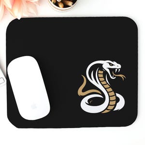 Puede incluir: Alfombrilla de ratón rectangular negra con un diseño de cobra blanca con detalles dorados y marrones. Un ratón de ordenador blanco está a la izquierda de la alfombrilla. La alfombrilla está diseñada para uso informático.