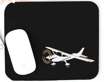 Puede incluir: Alfombrilla de ratón rectangular negra con un gráfico de avión blanco y detalles dorados. Un ratón Apple blanco está a la izquierda. La alfombrilla está diseñada para uso informático y añade un toque de aviación a cualquier espacio de trabajo.