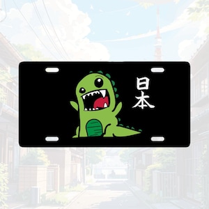 May include: A rectangular black license plate featuring a cartoon green dinosaur with a wide open mouth and sharp teeth. To the right of the dinosaur, the Japanese kanji characters for Japan, 日本, are printed in white. The plate has four mounting holes.