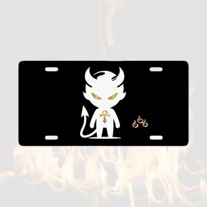 May include: A rectangular black number plate featuring a white cartoon devil character with gold eyes and a gold ankh symbol on its chest. To the right of the character, the number 666 is printed in a gold stylised font. The plate has four mounting holes.