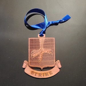 May include: A wooden ornament with a brown woodgrain finish. The ornament features a detailed laser-cut design of a bird's talon with the word "STRIKE" inscribed on a banner below. The ornament is attached to a blue ribbon.